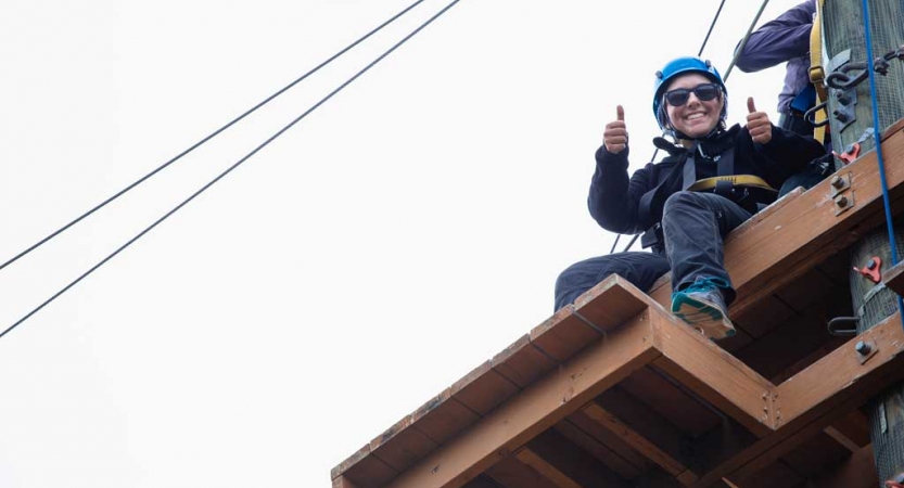 A person wearing safety gear and secured by ropes sits atop a wooden structure of a high ropes course, looks down and gives the camera two thumbs up.